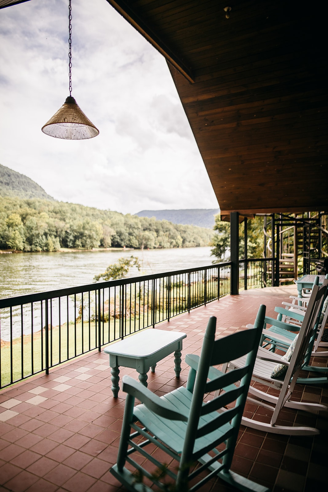 A spacious wrap-around porch features several rocking chairs in a calming teal hue. A view of the Tennessee River can be seen in the background, surrounded by lush greenery and distant mountains under a partly cloudy sky.