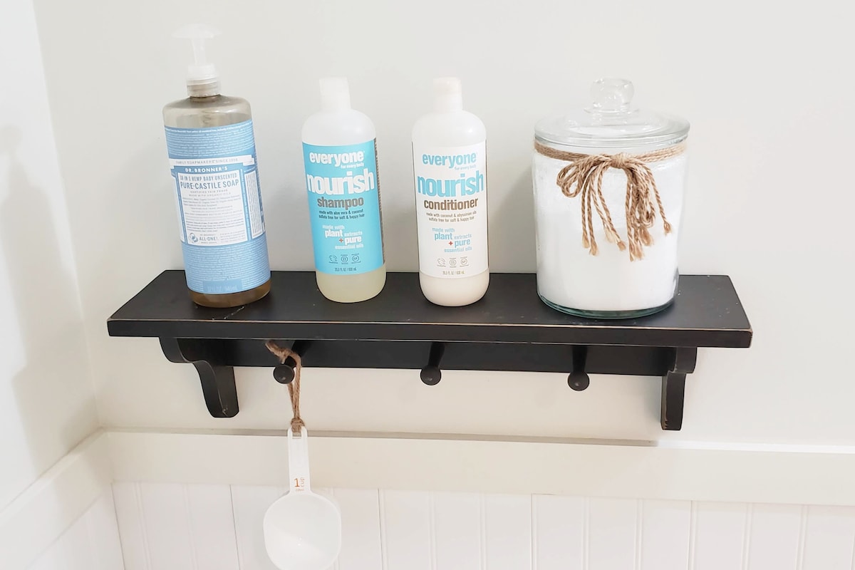 A wall-mounted shelf displays four bath products: a bottle of body wash, two bottles of shampoo and conditioner, and a glass jar with a lid tied with twine. A small measuring scoop hangs from the shelf, adding functionality.