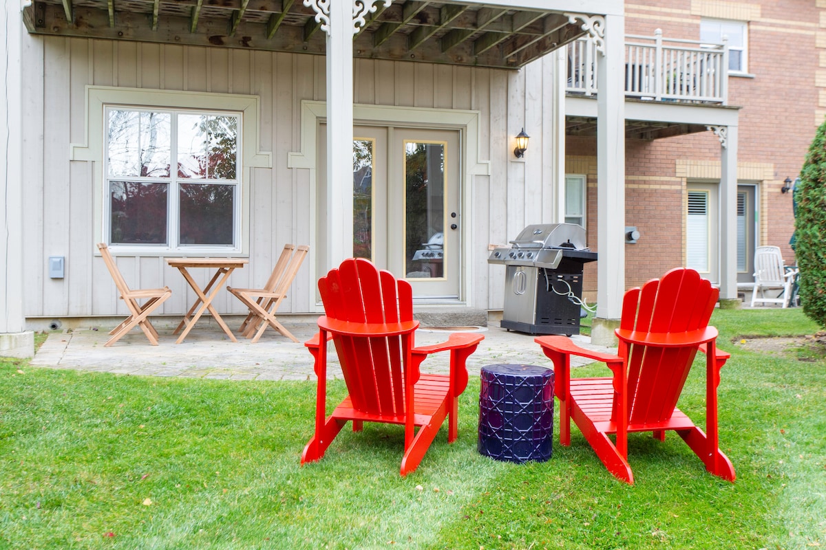 A back patio is featured, showcasing two red Muskoka chairs positioned on a grassy area. A small round table is seen between them, accompanied by a gas barbecue grill. Large glass doors provide access to the chalet's interior, with a portion of the building visible in the background.