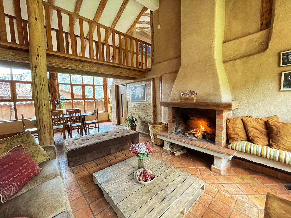 A cozy living area features a rustic fireplace with a warm fire, surrounded by comfortable seating. Natural light enters through large windows, highlighting the wooden beams and terracotta floor tiles. A dining area is visible in the background, creating a welcoming open space.