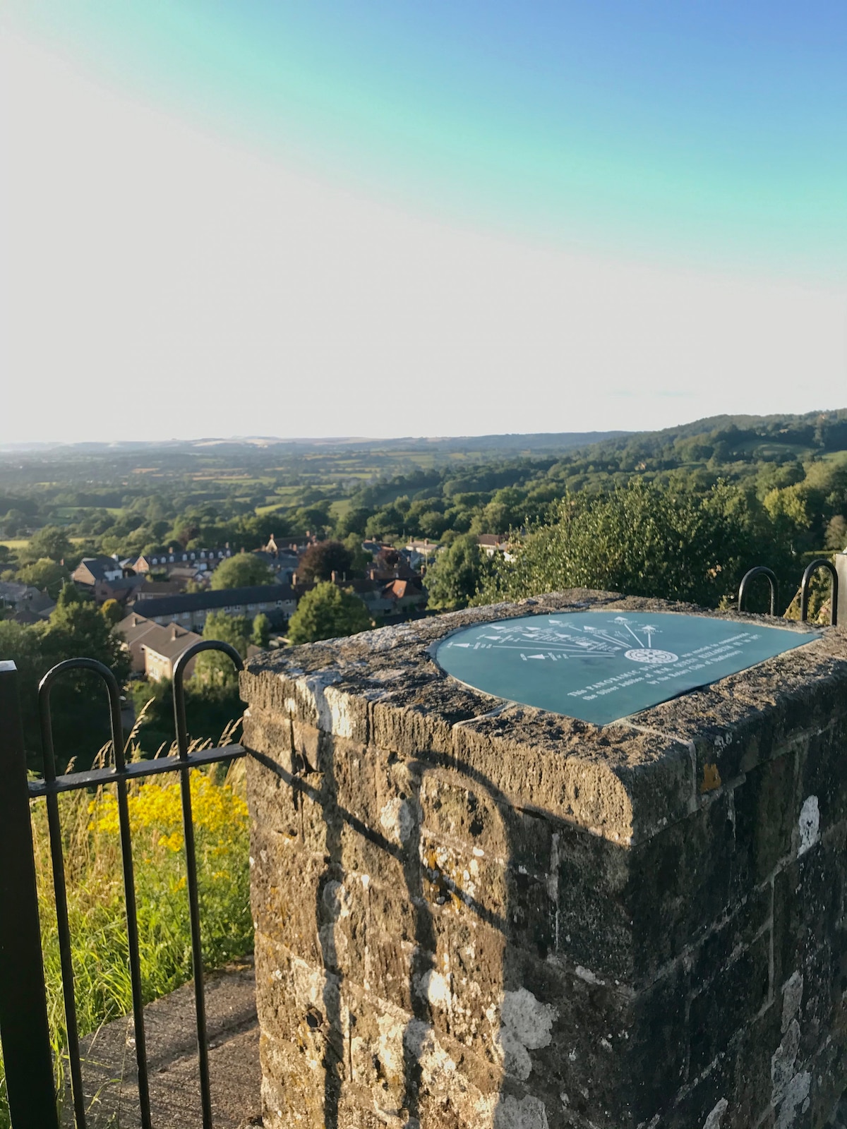 A stone marker is positioned at the edge of a viewpoint, surrounded by a wrought iron fence. The landscape beyond showcases rolling hills and a quaint village nestled in the valley, under a sky transitioning to evening light.