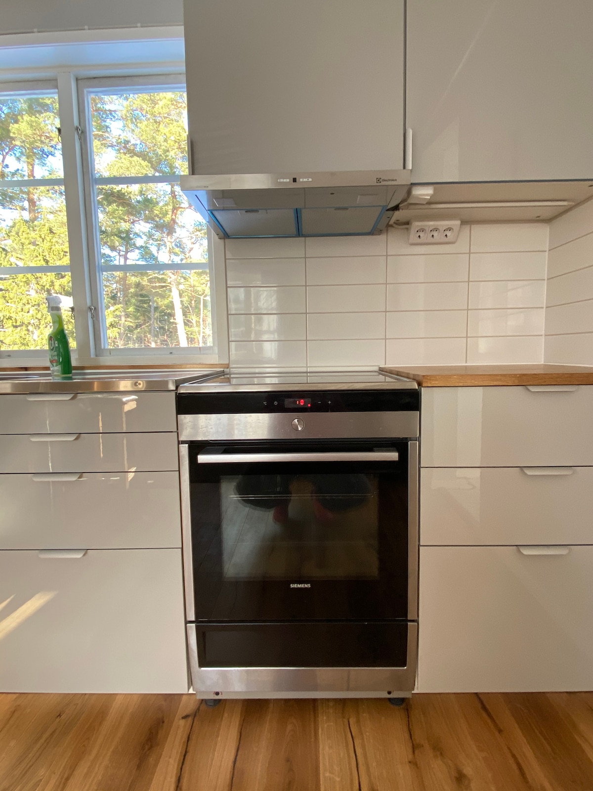 A modern kitchen is featured with sleek, white cabinetry and a minimalist design. A built-in oven with a black front is positioned centrally, complemented by a stainless steel extractor hood above. Natural light filters in through a large window, highlighting the warm wooden flooring.