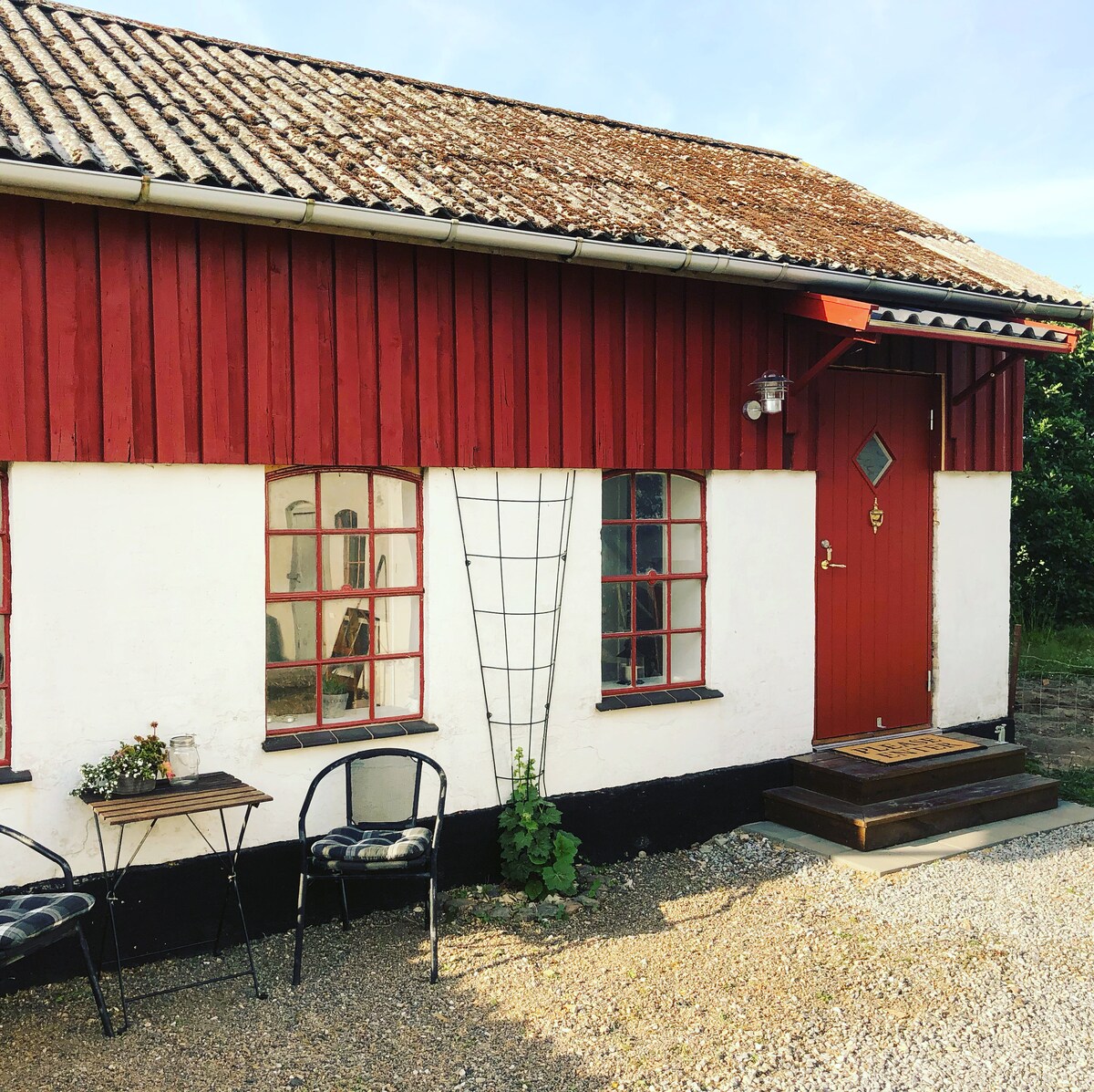 The exterior of a charming red building features a wooden facade and a slanted roof. A small set of steps leads to the entrance door, which is adorned with a diamond-shaped window. Two black chairs and a small table are positioned outside, inviting relaxation.