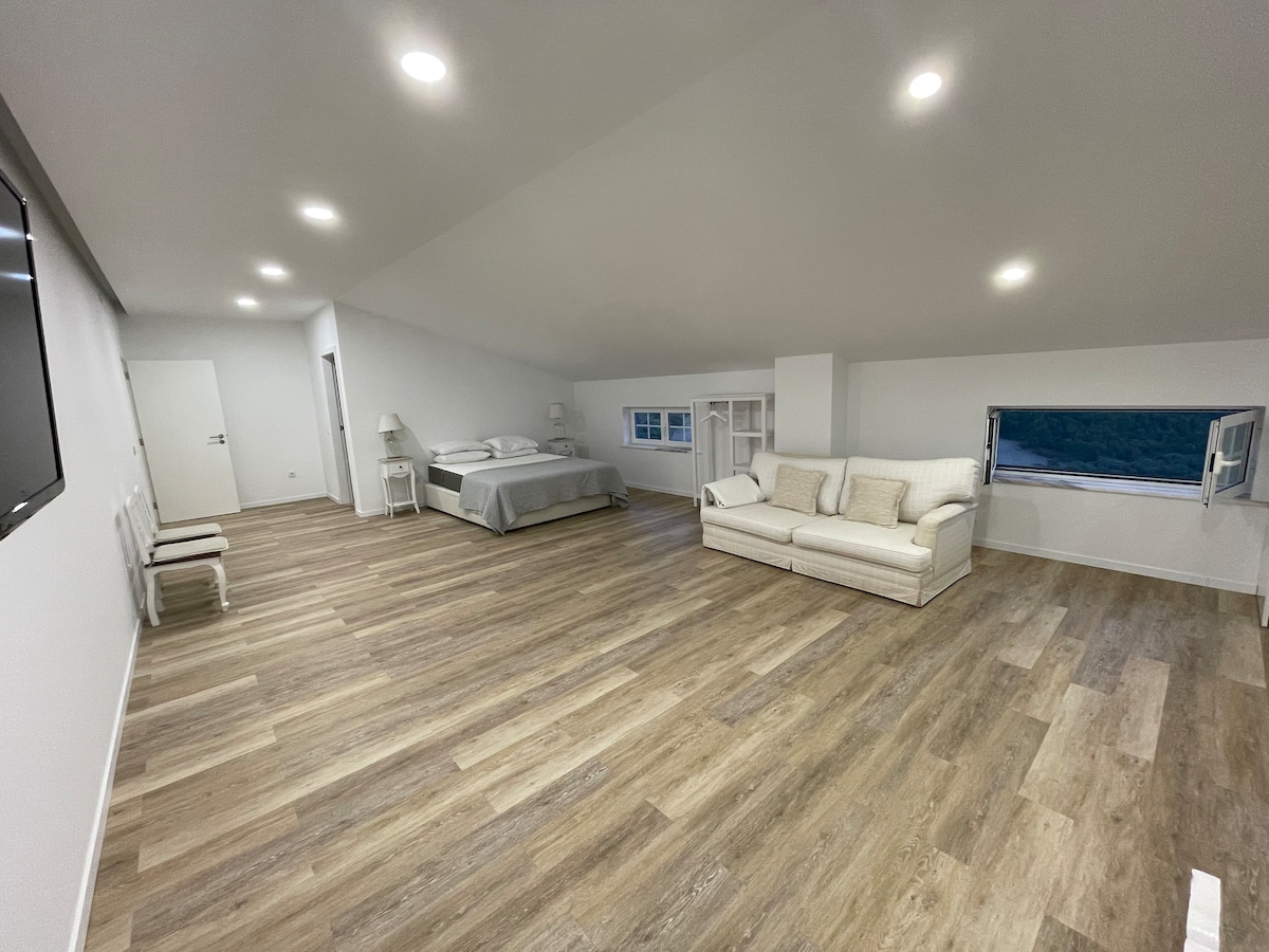 A spacious room is highlighted by light-colored wooden flooring and minimalist furnishings. A cozy sofa is positioned toward a window, while a large bed is set against the far wall. Soft ambient lighting enhances the open feeling of the space.
