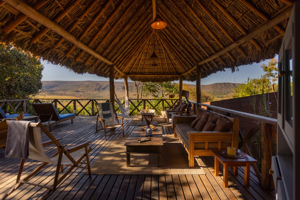 An open deck area features a thatched roof, surrounded by lush greenery. Several seating options, including wooden chairs and a table, provide comfortable spaces to relax. A scenic view of rolling hills can be seen in the background, enhancing the connection to nature.