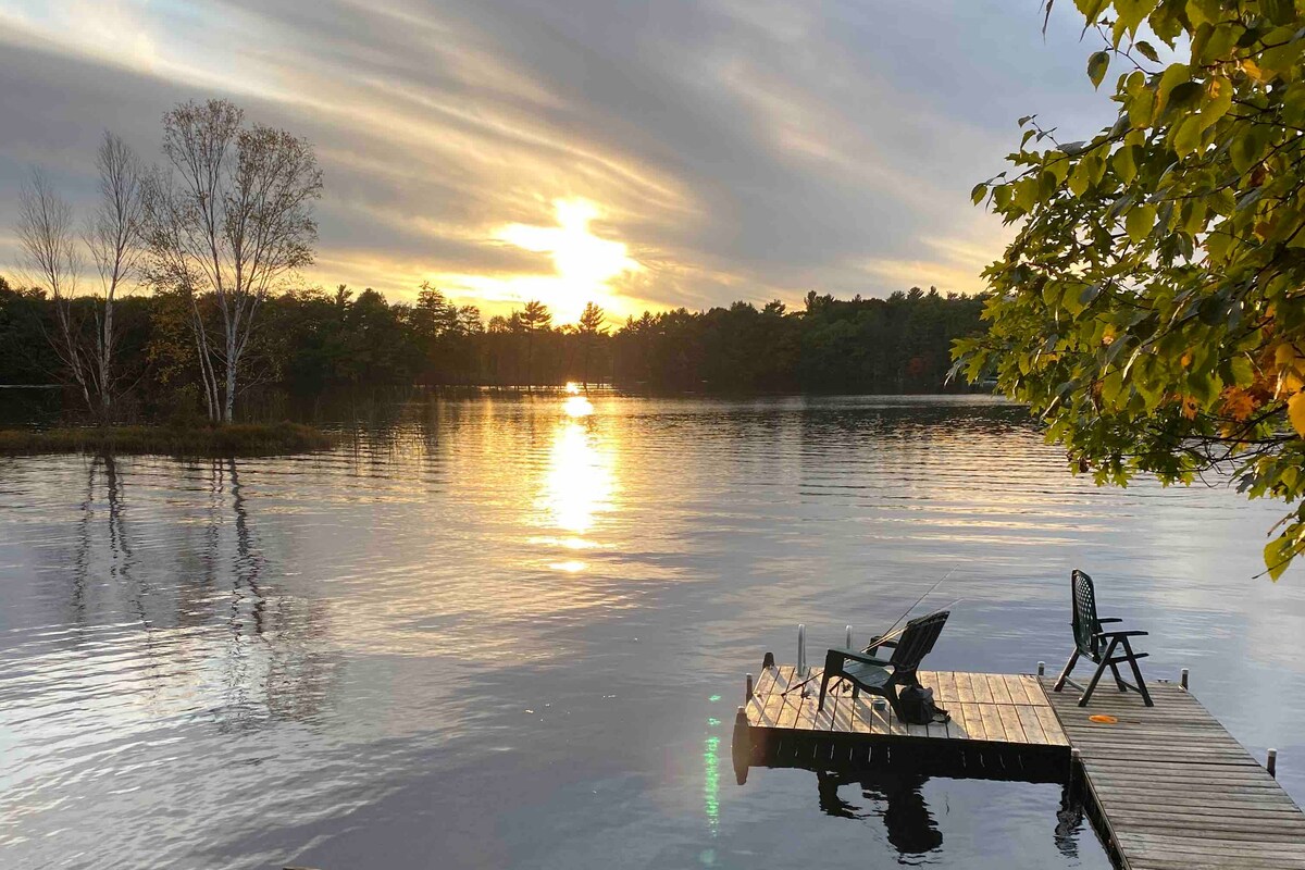 A serene lake view captures the setting sun above the calm water, casting reflections of warm colors. Two chairs are positioned on the wooden dock, inviting relaxation while surrounded by trees with colorful foliage.