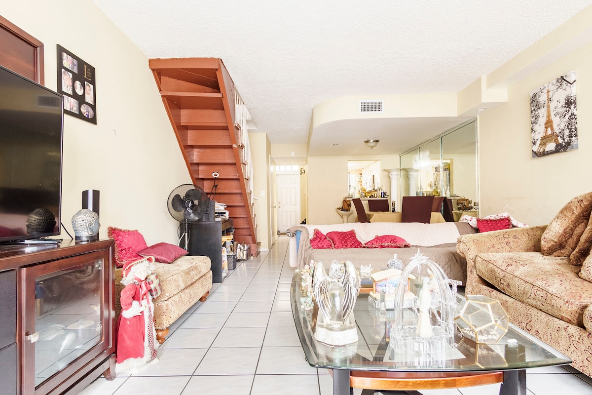 A spacious living area features light-colored tiles and a combination of sofas adorned with various cushions. A wooden staircase ascends to the upper level, while a glass coffee table showcases decorative items. Reflections are seen in the mirrored wall at the back.