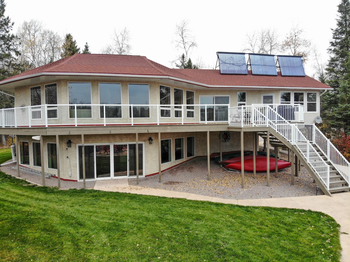 The modern two-level home features an octagonal design, with expansive glass doors and a spacious wrap-around deck. A set of stairs leads to the main entrance, while outdoor areas are surrounded by well-kept grass and trees, offering a serene natural setting.