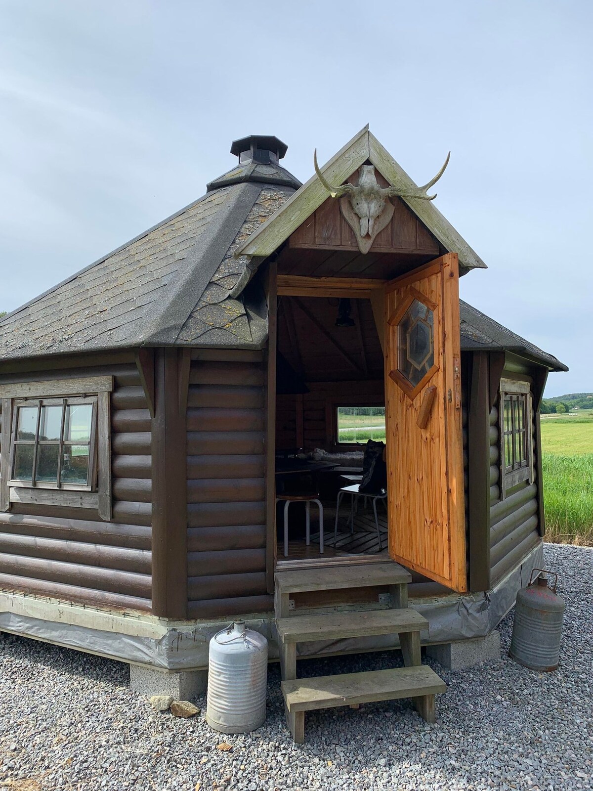 A rustic grill cabin features a wooden exterior with a triangular roof and a welcoming door. The entrance is flanked by large windows, providing natural light inside. The cabin is situated on a gravel area, with lush greenery visible in the background.