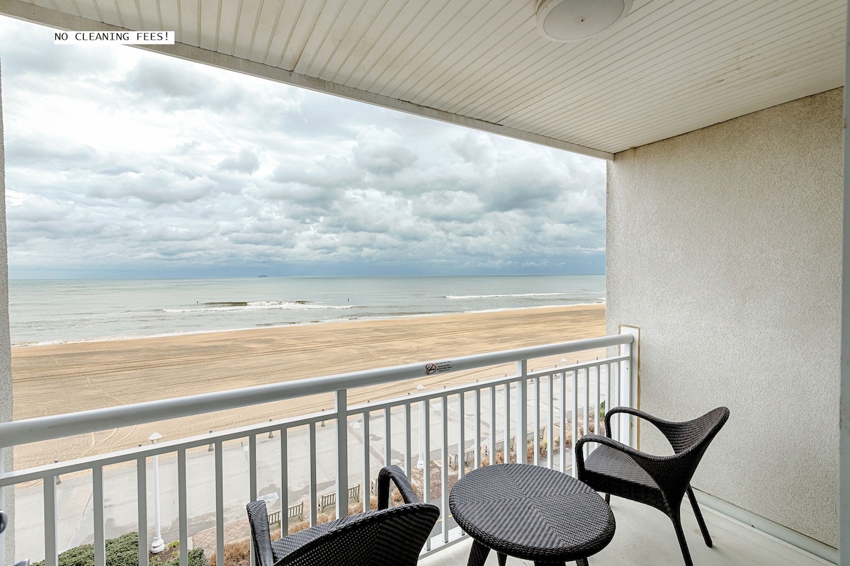 An oceanfront balcony features two wicker chairs paired with a small table. The expansive view showcases the sandy beach meeting the sea under a cloudy sky, with gentle waves visible in the distance.