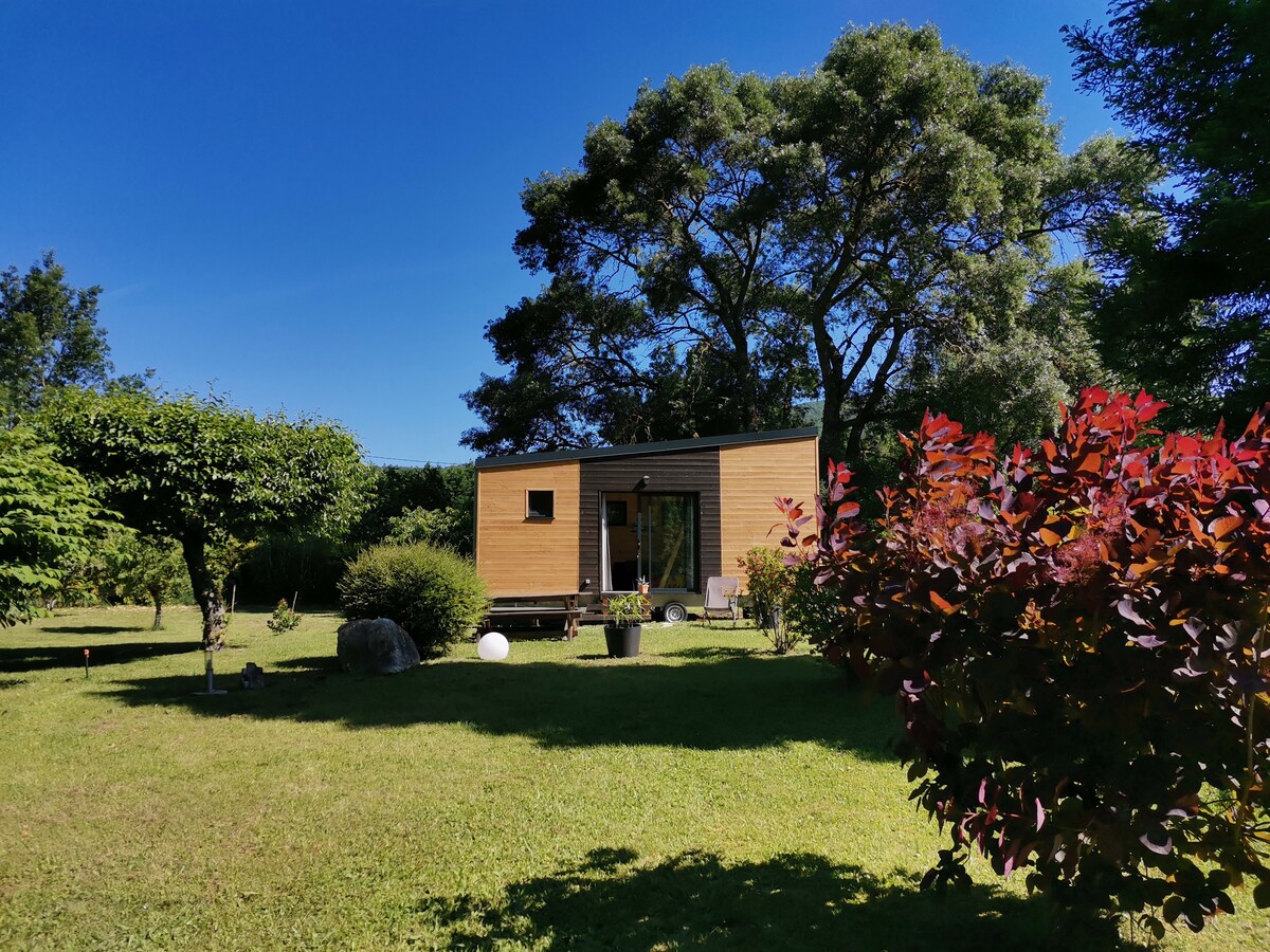 The tiny house is nestled in a well-kept garden, surrounded by lush greenery and flowering plants. A clear blue sky complements the wooden exterior. The peaceful outdoor setting includes various shrubs and trees, enhancing the tranquil atmosphere.