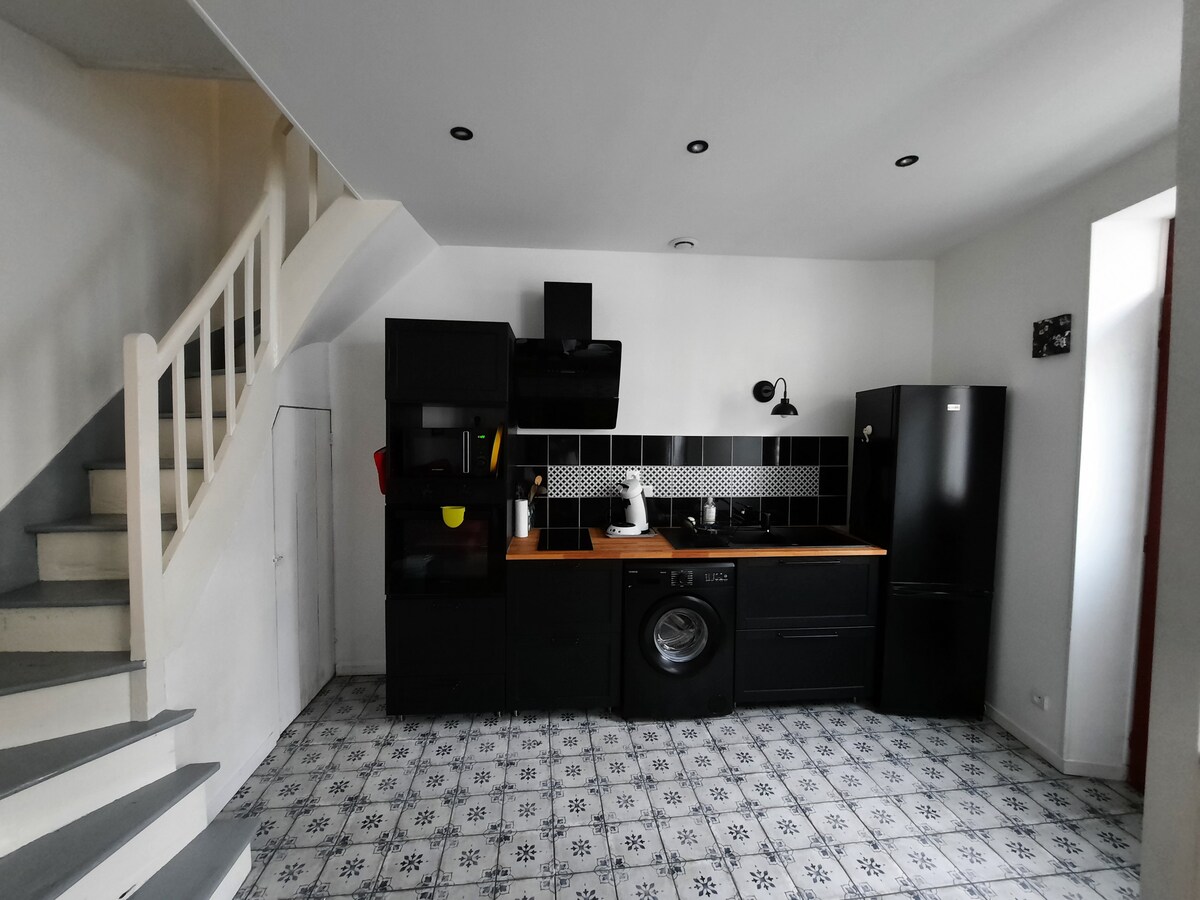 A modern kitchen area features black cabinetry with a wooden countertop. Appliances include a refrigerator, washing machine, and stove. Decorative black tiles are highlighted on the wall. A staircase is visible in the background, leading to the upper level.