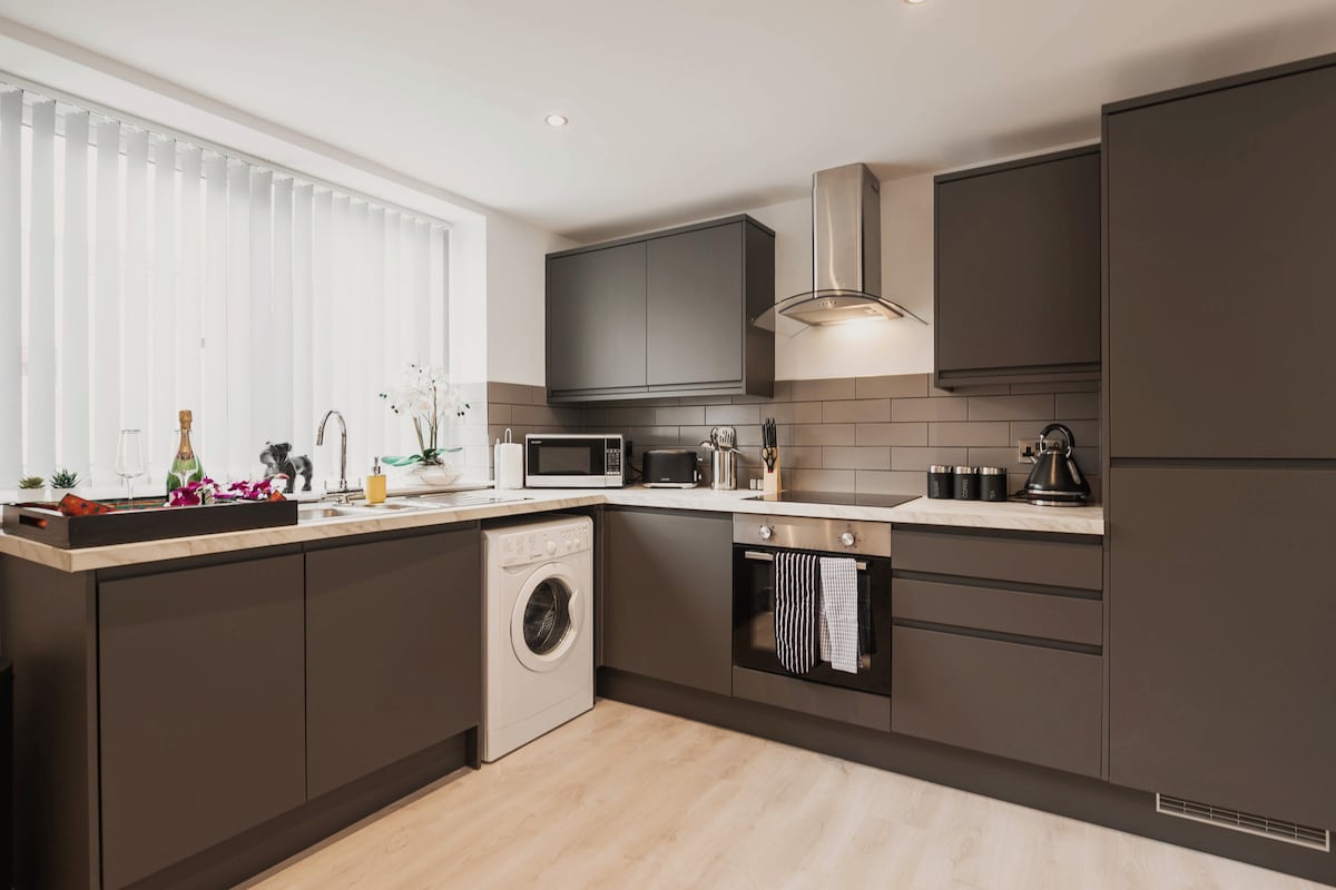 A modern kitchen is equipped with dark cabinetry and a light-colored countertop. Stainless steel appliances include a built-in oven, kettle, and microwave. A washer is integrated into the cabinetry. Vertical blinds allow natural light to enter, while a selection of kitchen essentials is neatly arranged.
