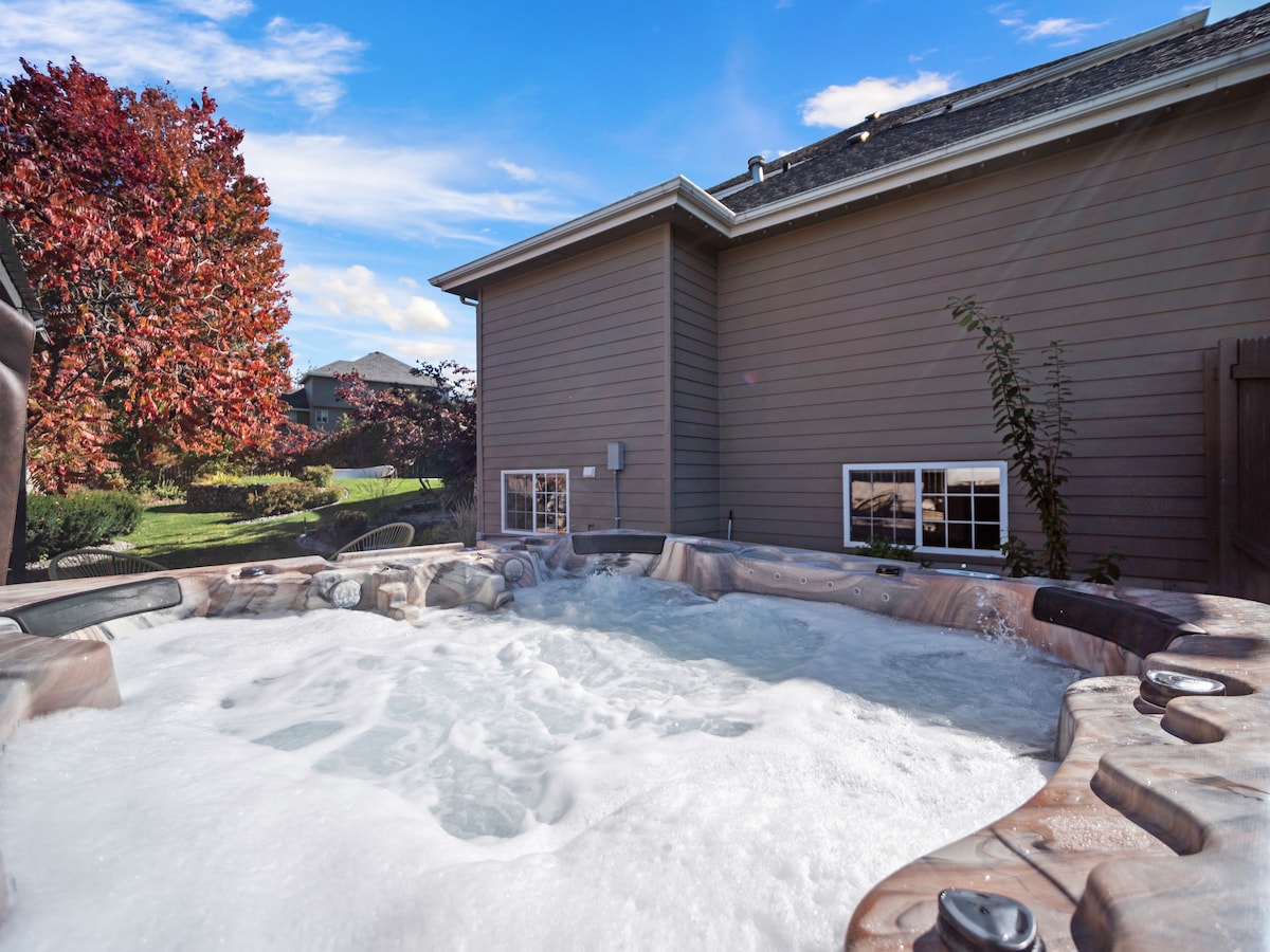A new hot tub is shown, surrounded by a wooden deck and positioned next to the home. Lush greenery and mature trees provide a backdrop, with bright blue skies overhead creating a serene outdoor setting.
