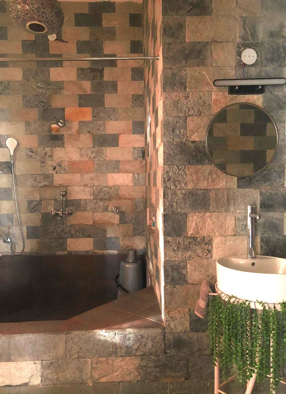 A spacious private bathroom is featured, showcasing textured stone walls in muted tones. The bathroom includes a large bathtub, a modern sink with a round mirror above, and a shower area with dual fixtures. Greenery adorns the sink area, enhancing the tranquil setting.
