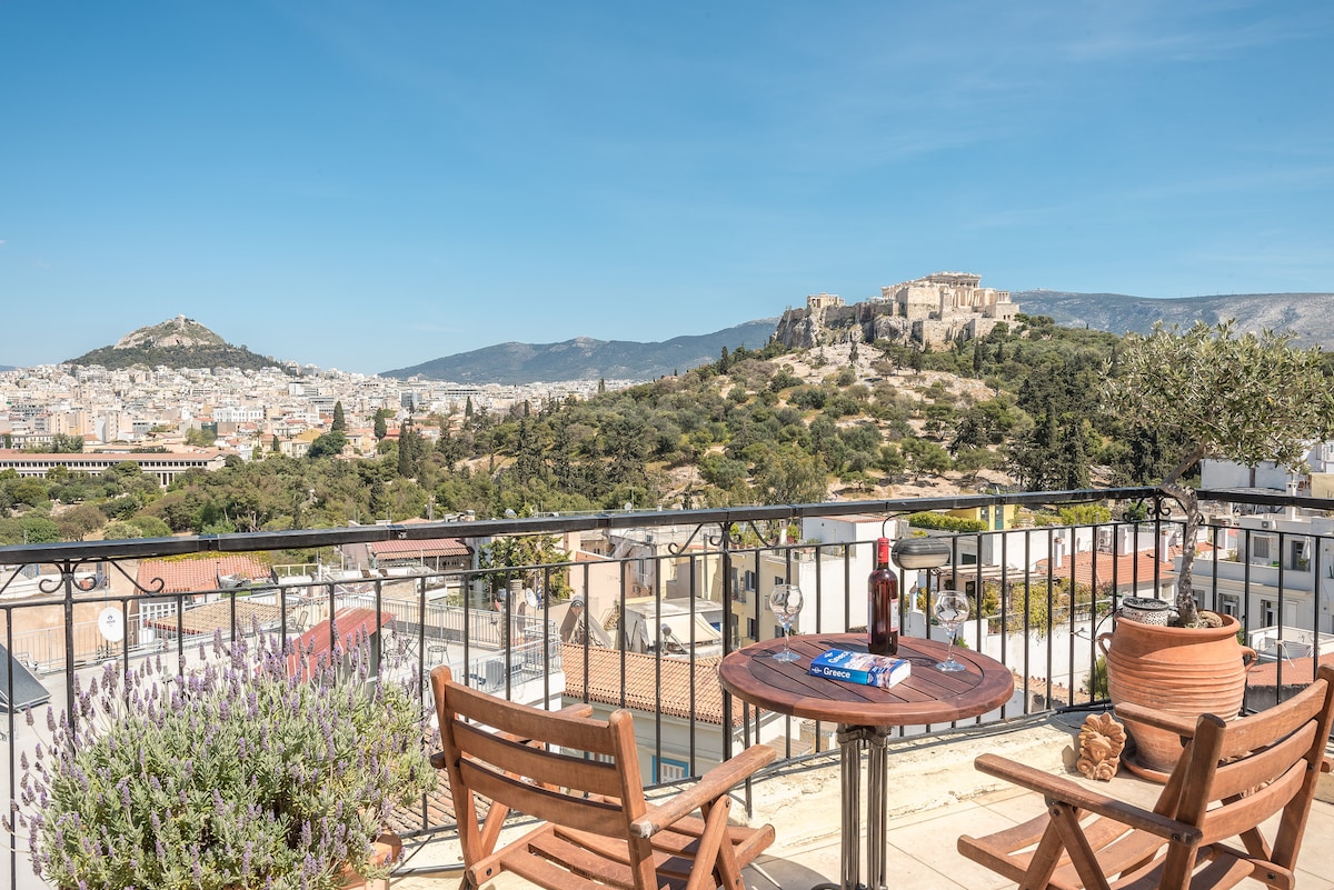 A shared rooftop terrace is presented with a wooden table and two chairs, offering a panoramic view of Athens, including the Acropolis and surrounding hills. Potted plants add greenery to the setting, while the clear blue sky enhances the overall ambiance.