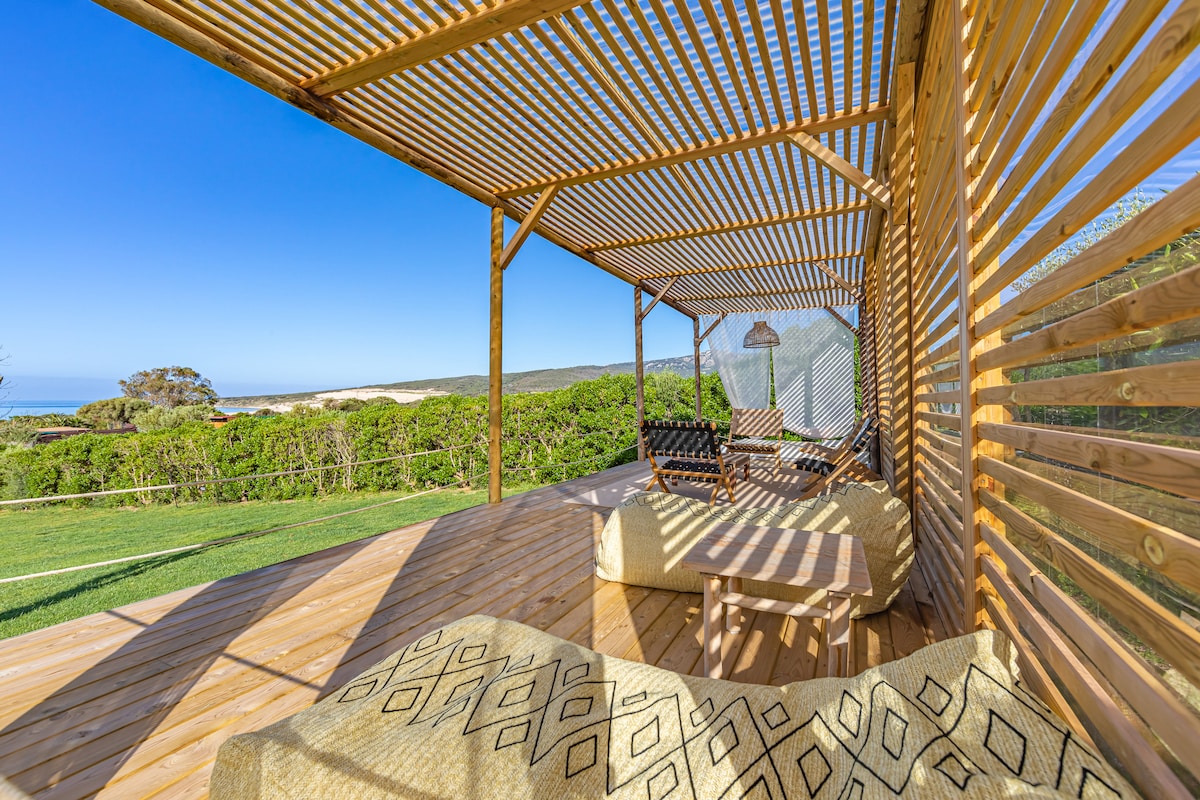 A spacious outdoor patio features wooden slats creating a shaded lounge area. Comfortable seating is arranged, offering views of the lush landscape and distant sea horizon. Natural light casts soft shadows on the wooden flooring, enhancing the tranquil atmosphere.