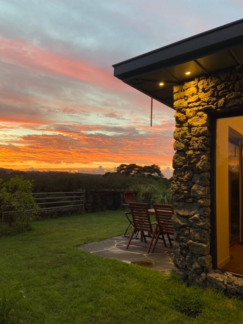 A stone structure with warm lighting displays a terrace set with a wooden dining table and chairs. Lush green grass surrounds the area, complemented by a vivid sunset sky filled with orange and pink hues.