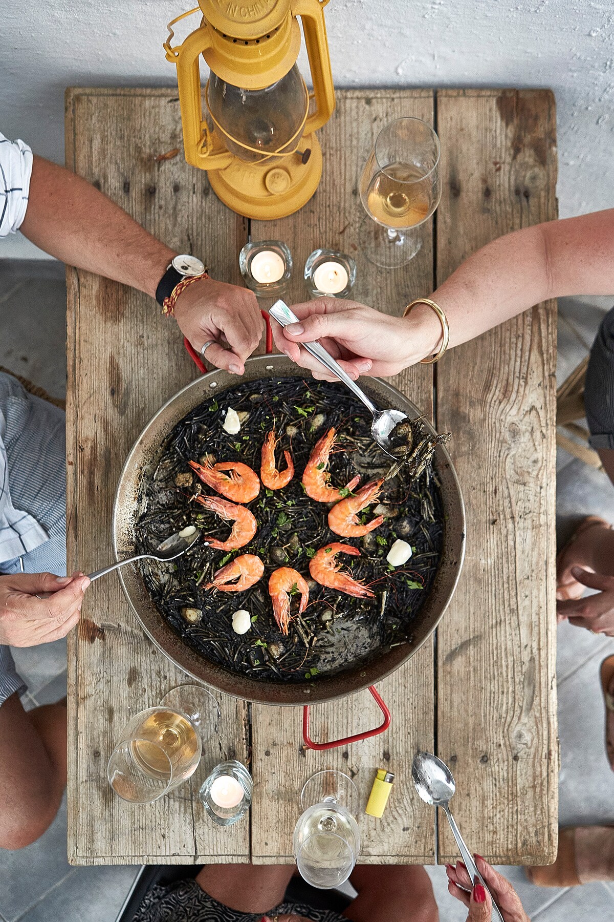A wooden table is set with a paella dish at its center, featuring vibrant shrimp arranged on a bed of dark rice. Two pairs of hands hold utensils, ready to serve. Glasses of white wine and candles add a warm ambiance.