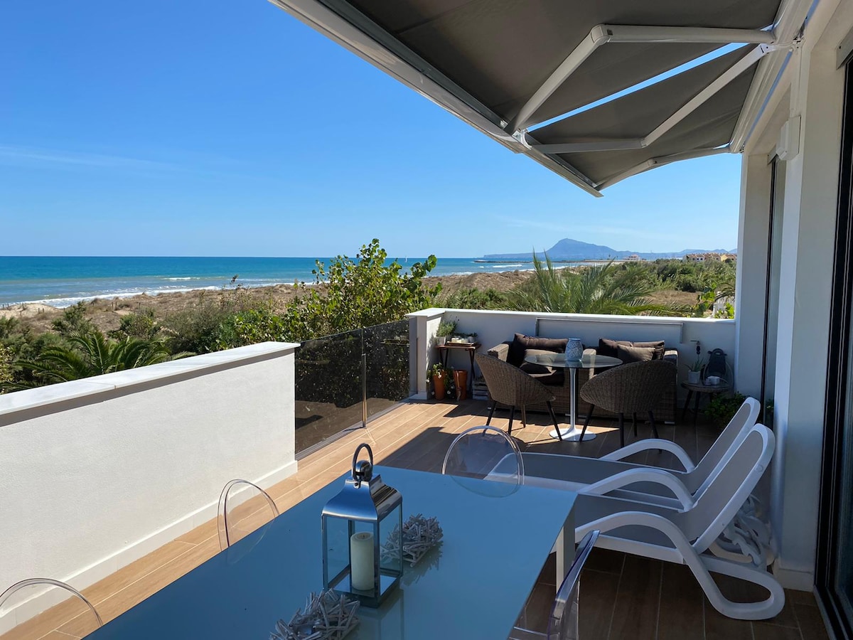A spacious terrace is shown overlooking a serene beach and ocean view. Outdoor dining furniture is arranged for relaxation, complemented by green plants nearby. A sunshade offers protection from the sunlight, creating a comfortable outdoor space for gathering.