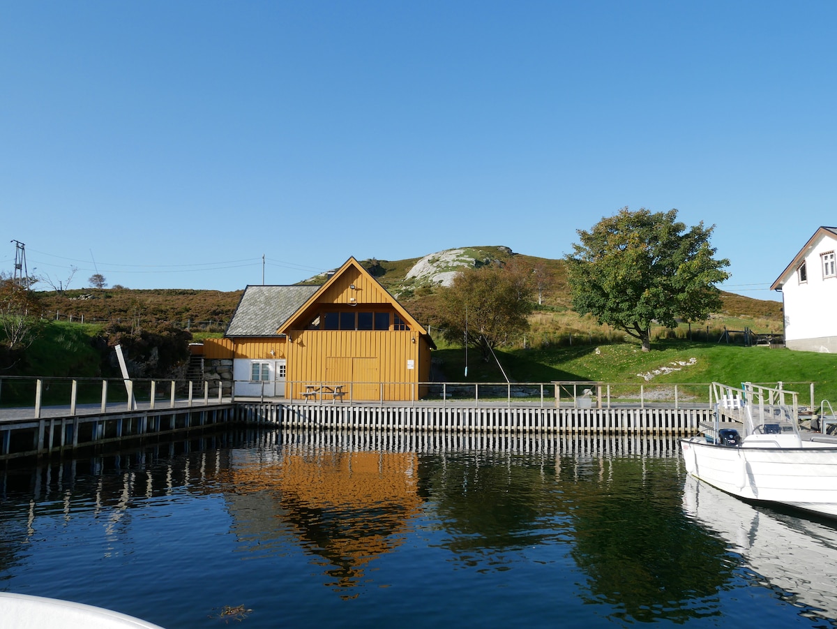 A charming yellow house is situated by a serene body of water, with its reflection visible on the surface. Lush greenery and a hillside are seen in the background, along with a dock and a couple of boats moored nearby.