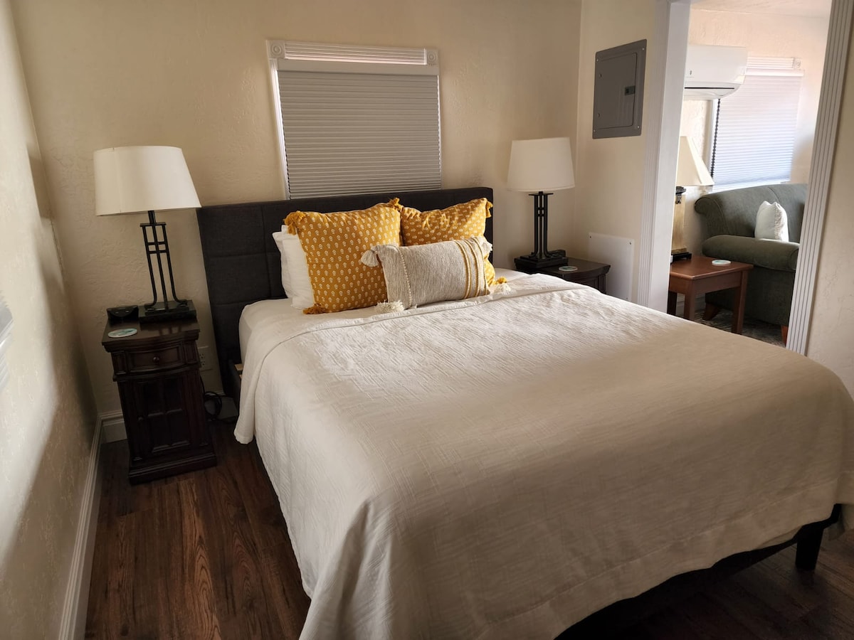 A comfortable bedroom features a spacious bed with a light quilt and decorative yellow pillows. Two bedside tables with lamps flank the bed. Natural light enters through a window, creating a bright atmosphere. A cozy seating area is subtly visible in the background.
