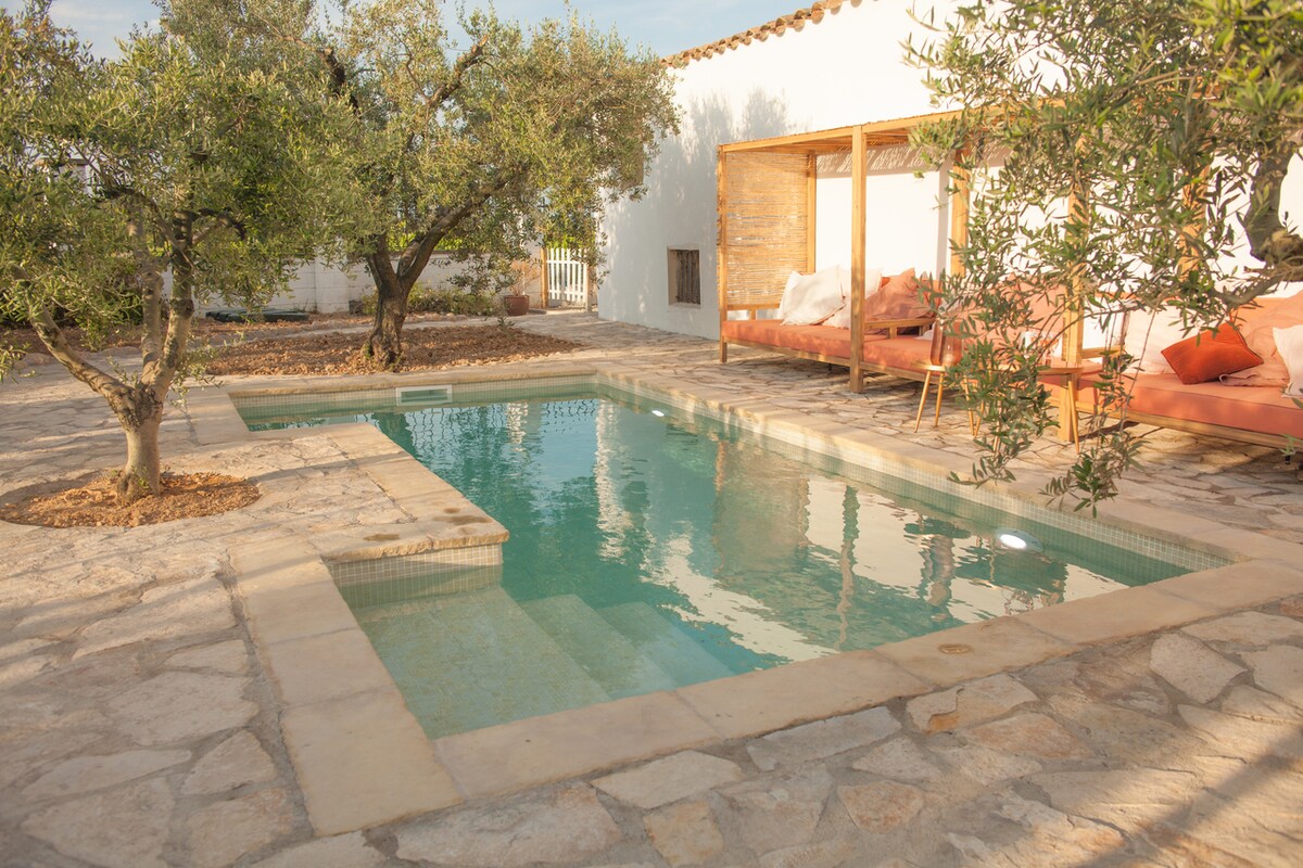 A serene outdoor pool is surrounded by natural stone paving, complemented by olive trees. A wooden pergola provides shade over lounge seating, creating a tranquil retreat atmosphere. The clear water reflects the surrounding greenery and provides a inviting spot for relaxation.