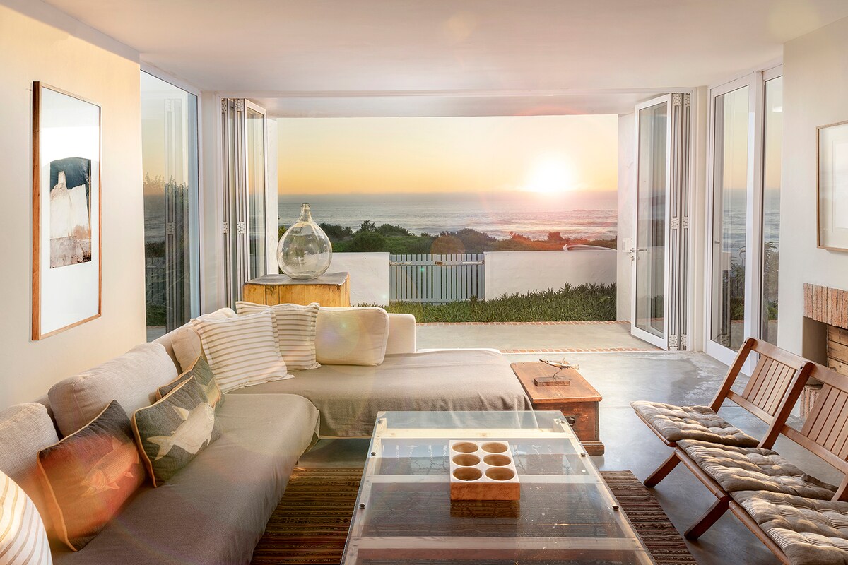 A bright living area features expansive glass doors that open to reveal a view of the ocean at sunset. A comfortable sofa, wooden coffee table, and stylish chairs are arranged to facilitate conversation and enjoyment of the surroundings.