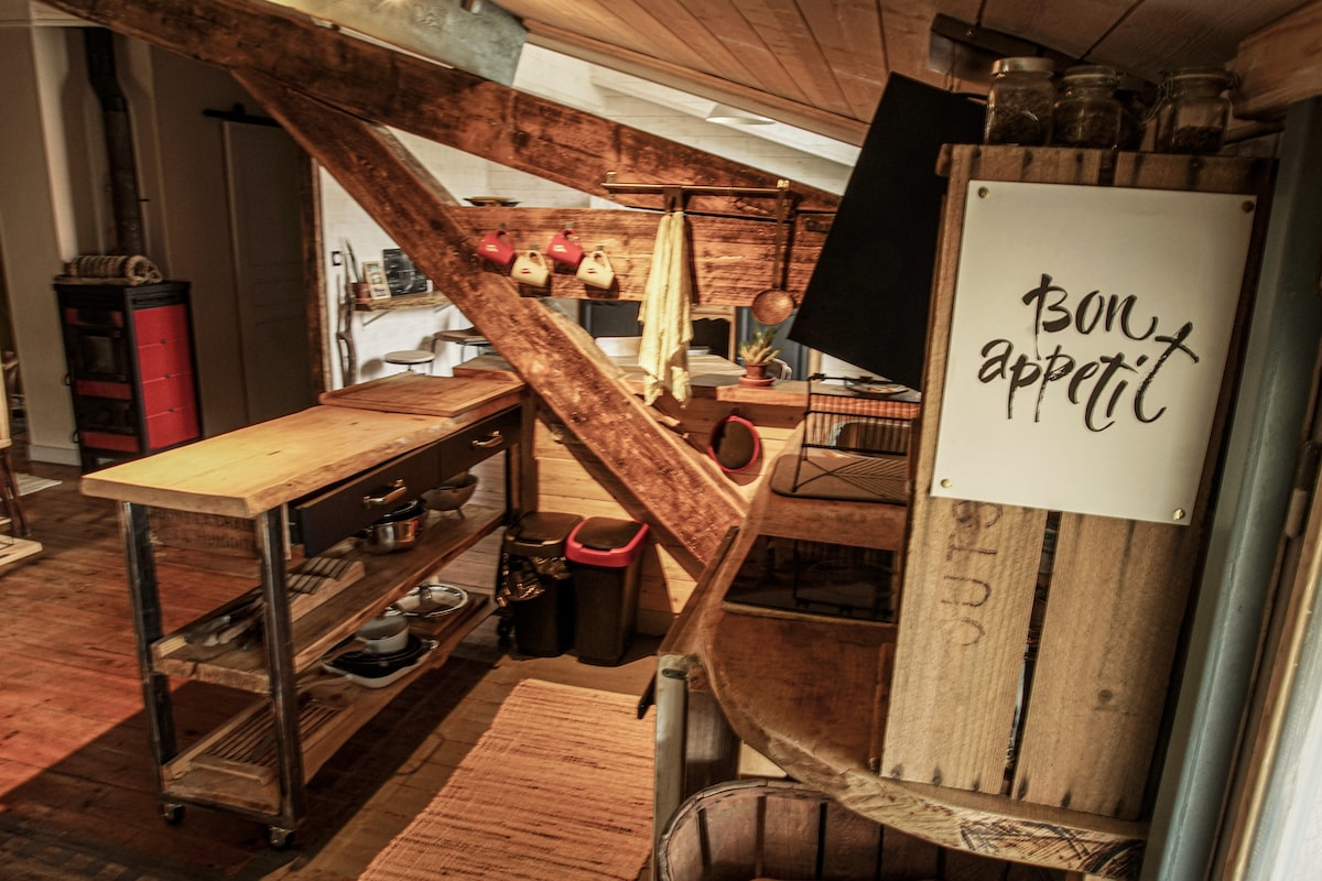 The kitchen area features a combination of wood and metal elements, showcasing open shelving and storage solutions. A sign with 'Bon appétit' hangs above a wooden countertop, creating a welcoming environment. Natural light filters through the space, enhancing the rustic decor and thoughtful layout.