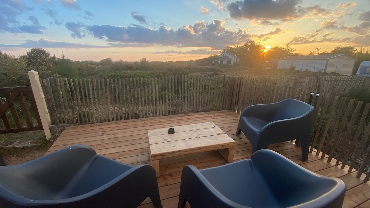 A wooden deck features three dark lounge chairs surrounding a rustic coffee table. In the background, the sun sets over a serene landscape bordered by natural vegetation, casting a warm glow over the scene.