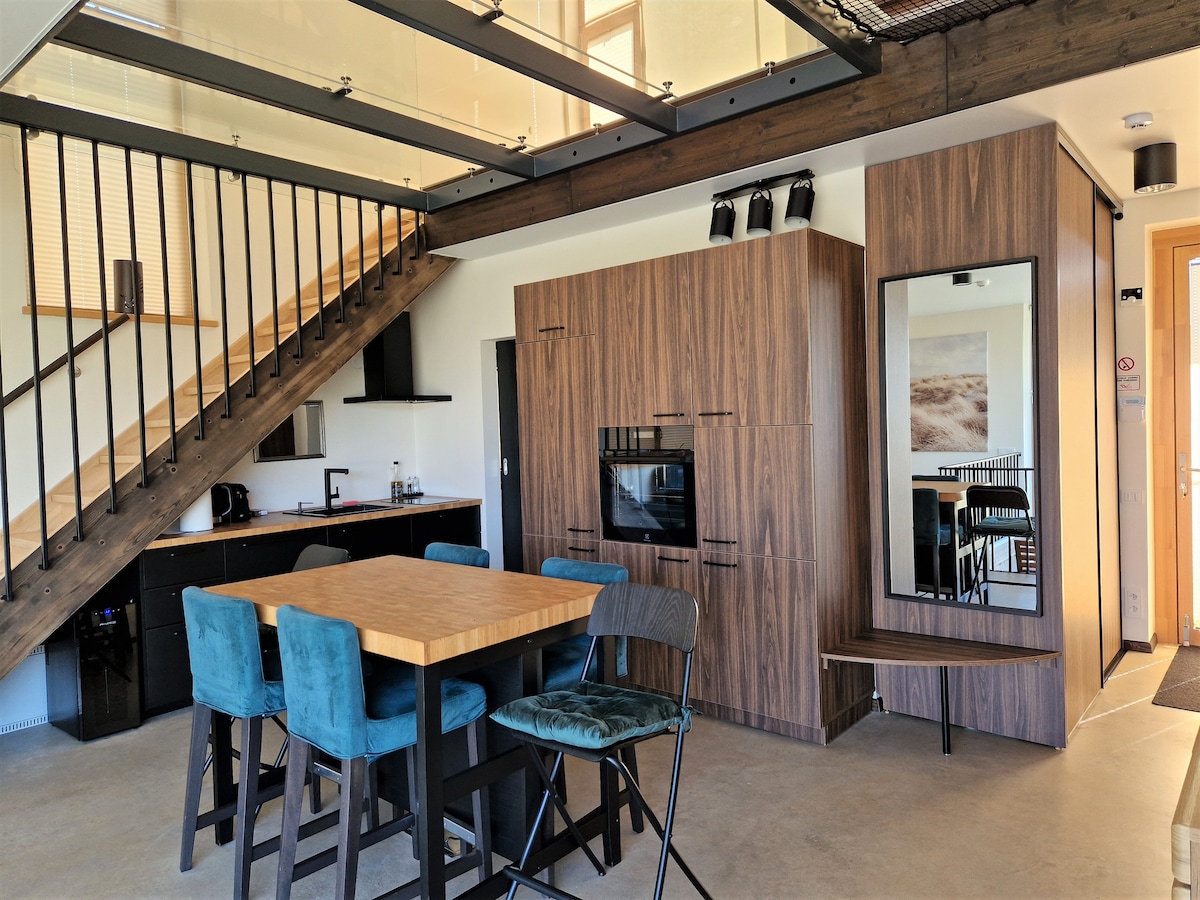 An open living area features a wooden dining table surrounded by upholstered chairs. A modern kitchen is visible in the background, complemented by stylish cabinetry. Natural light floods the space through large windows, enhancing the minimalistic design and inviting atmosphere.