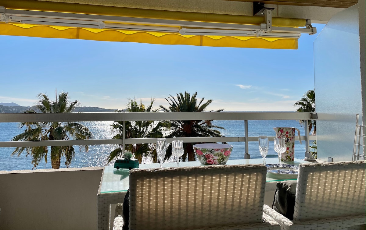 A terrace features a dining table set for four, complemented by palm trees in view and the bay of Bandol in the background. Sunlight reflects off the water, and a bright yellow awning adds a pop of color to the space, providing shade.