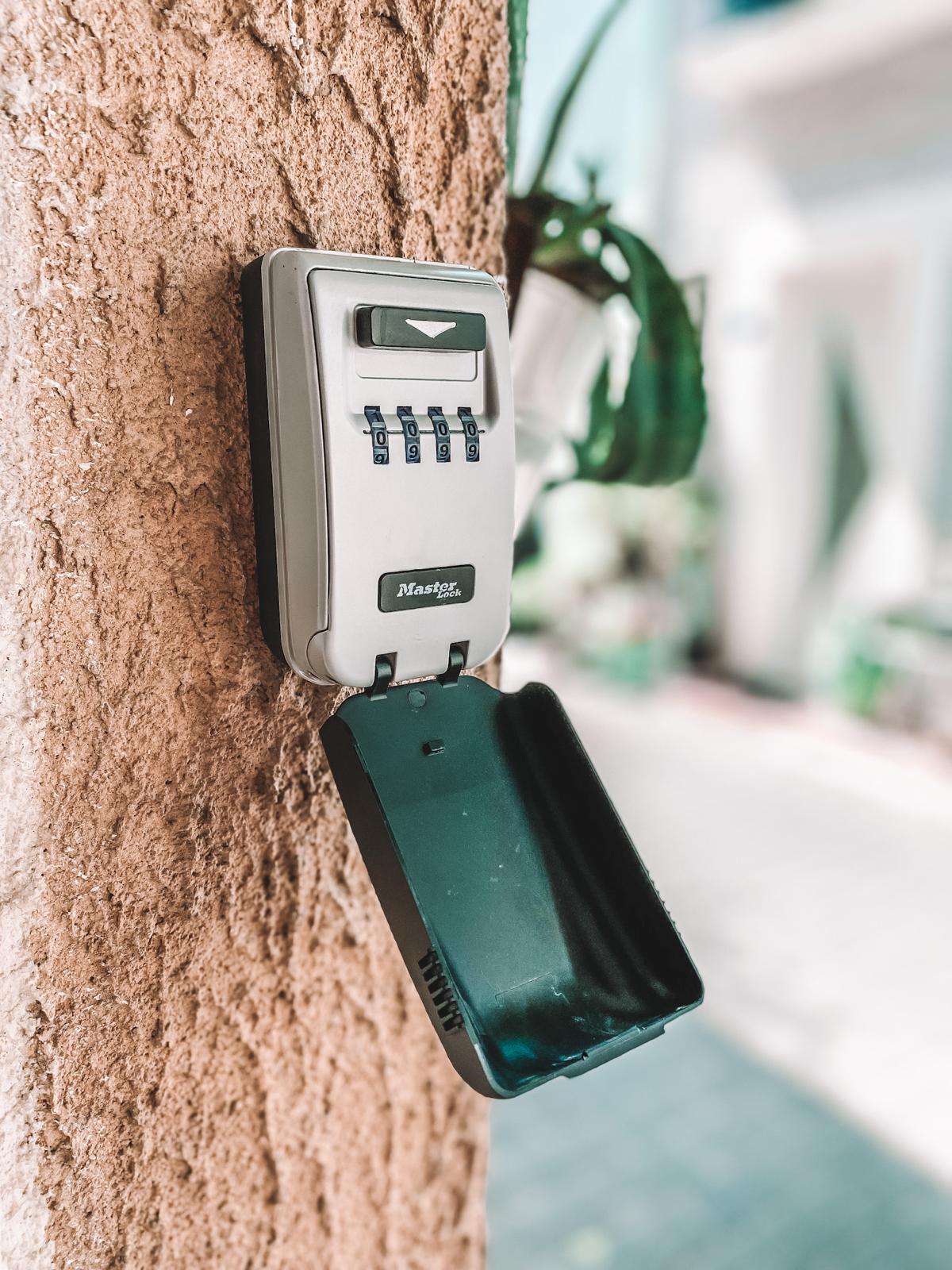A secure combination lockbox is mounted on a textured wall. The lockbox is opened to reveal a black compartment for key storage. Surrounding greenery and blurred background elements suggest a well-maintained outdoor area.