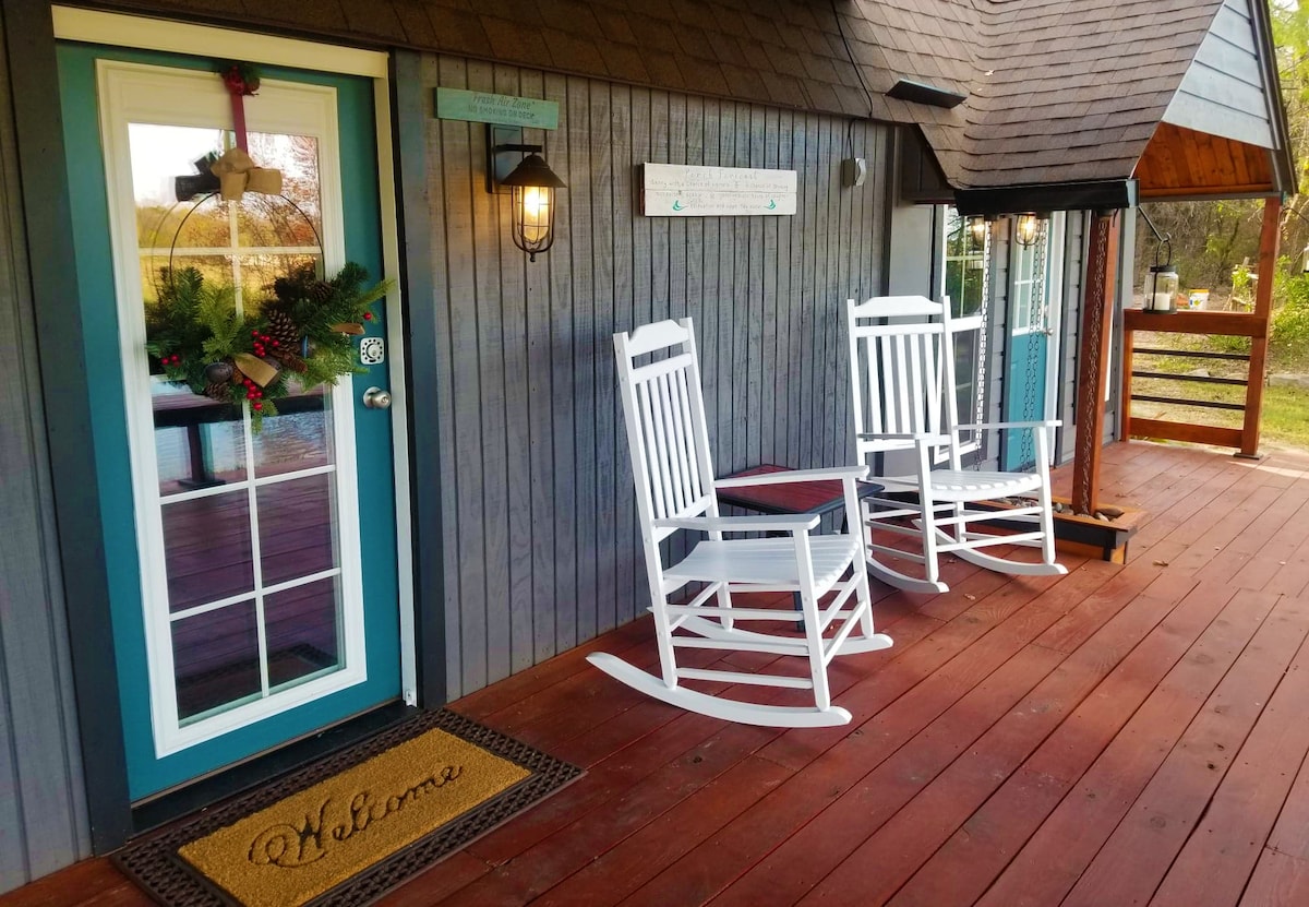 The entrance features a welcoming door adorned with seasonal decor. Two white rocking chairs are positioned on the covered deck, complementing the wooden flooring. A 'Welcome' mat is placed at the door, inviting guests to enter and enjoy the serene surroundings.