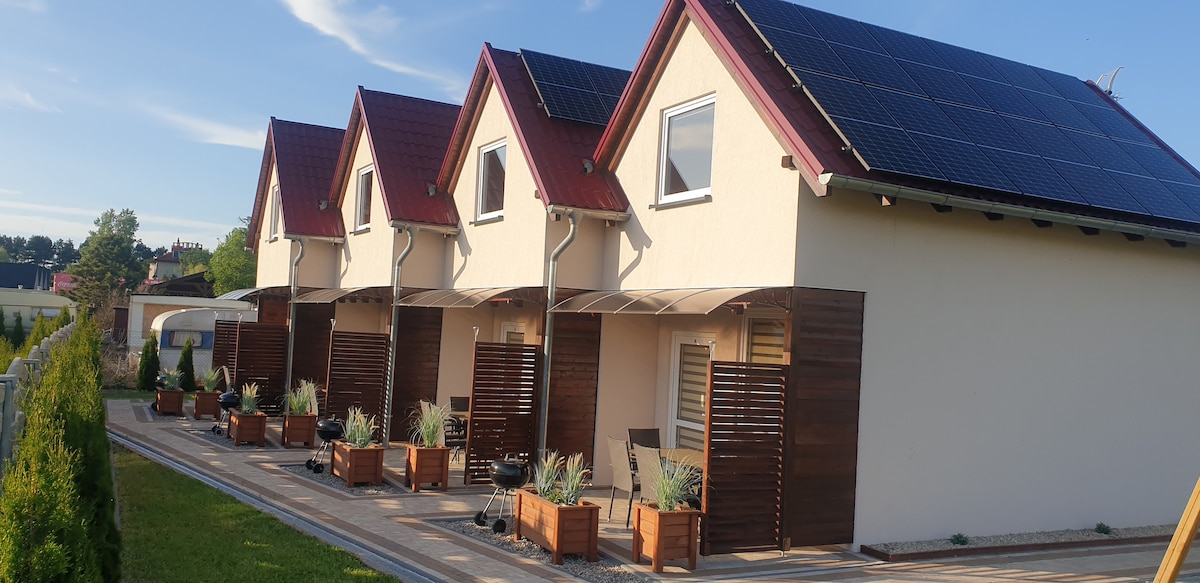 An arrangement of multiple cottages is visible, each equipped with solar panels on the roof. Wooden planters with greenery line the front, while shaded patios with seating areas offer outdoor relaxation. The design combines modern efficiency with inviting outdoor spaces.