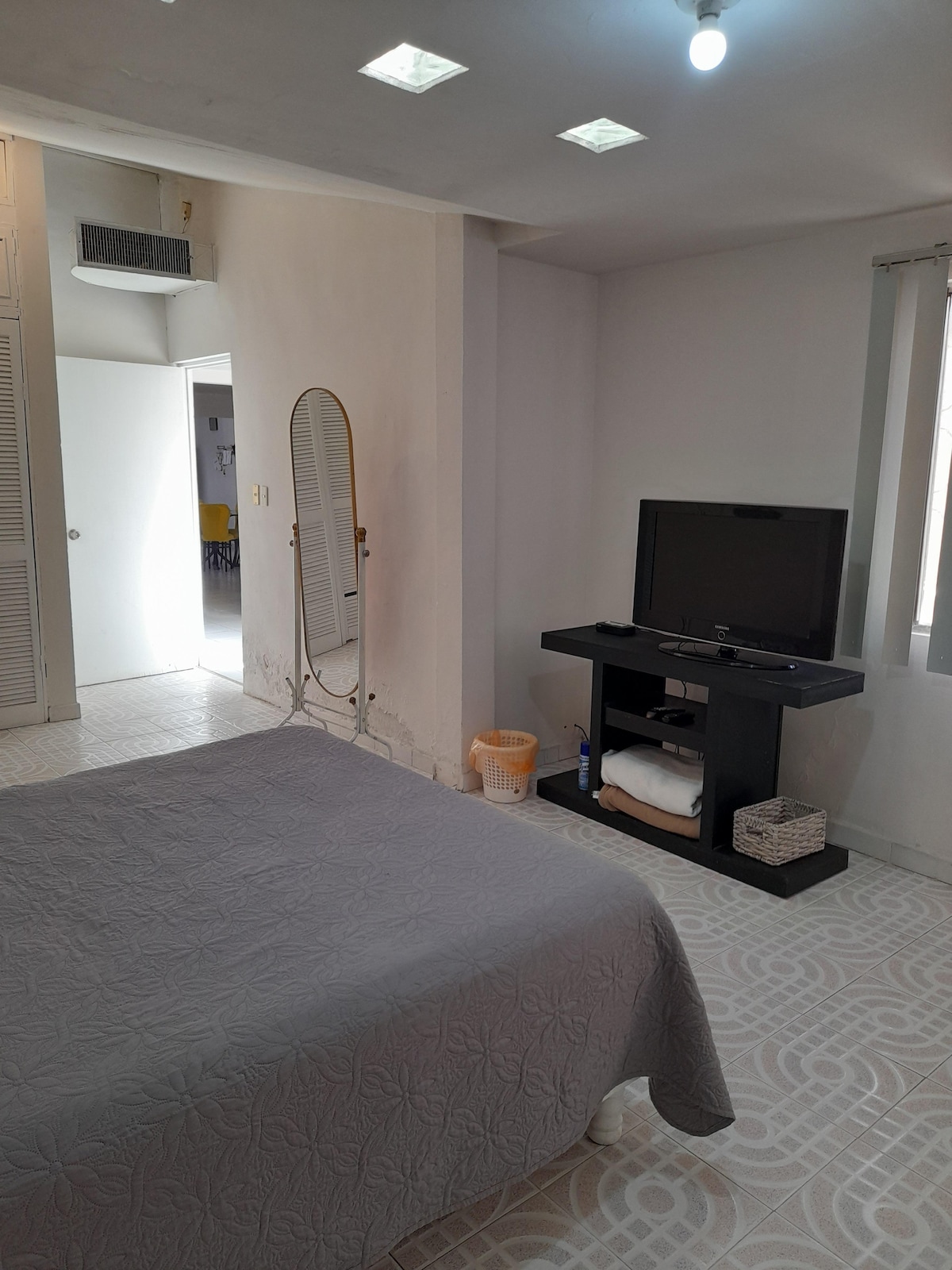 A spacious bedroom features a queen-size bed covered with a light gray quilt. A mirror and television are positioned against the wall. Natural light enters through the window, complemented by light-colored curtains. A wastebasket and towels are neatly arranged on a shelf nearby.