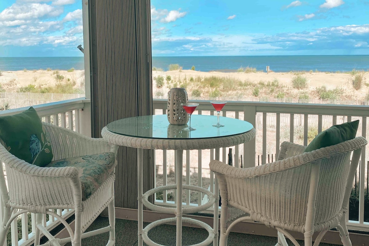 A round glass table is set on a private balcony, accompanied by two white wicker chairs. A candle holder and two glasses are positioned on the table, with a view of the sandy beach and ocean in the background. The sky is partly cloudy.