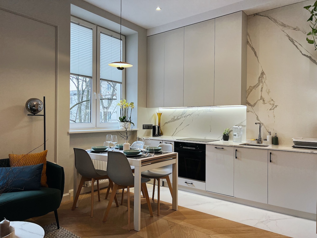 A modern kitchen area features sleek cabinetry with a marble backsplash, equipped with essential appliances. A dining table set for two is complemented by contemporary chairs. Natural light filters through the double windows, highlighting the warm tones of the wooden flooring.