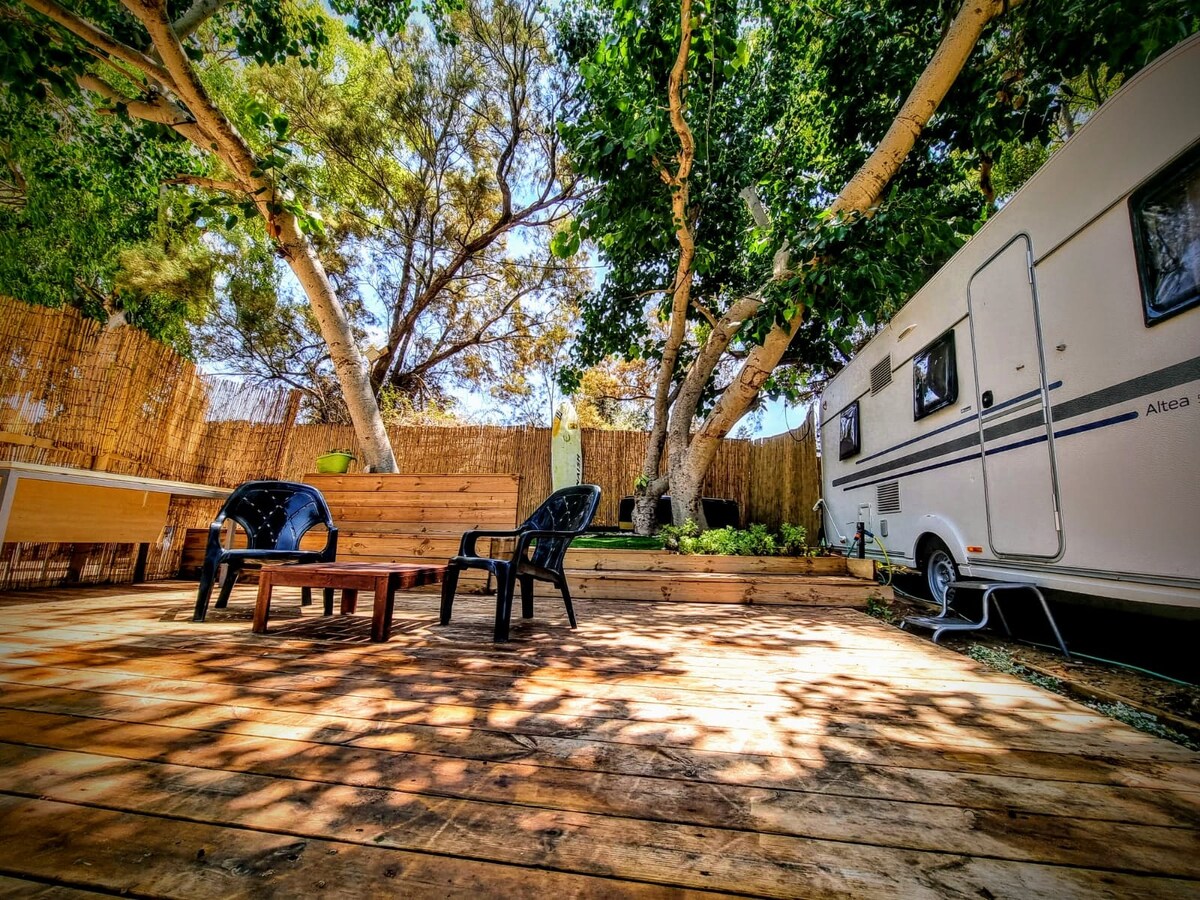 A wooden deck is surrounded by lush greenery, featuring a small table and several chairs. A caravan is parked nearby, partially shaded by large trees. Dappled sunlight creates patterns on the deck floor, contributing to the natural ambiance of the space.