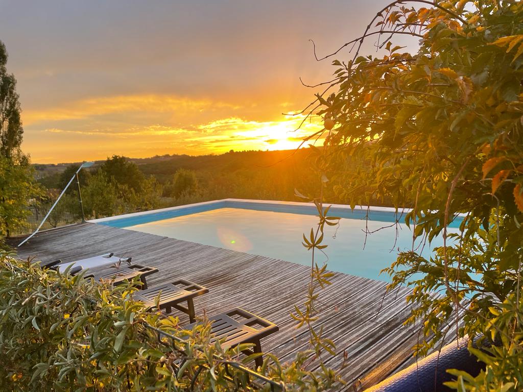 The image captures a serene swimming pool surrounded by wooden decking. Sunlight reflects off the water's surface, with lush greenery framing the scene. A stunning sunset fills the background, casting warm colors across the sky and landscape.