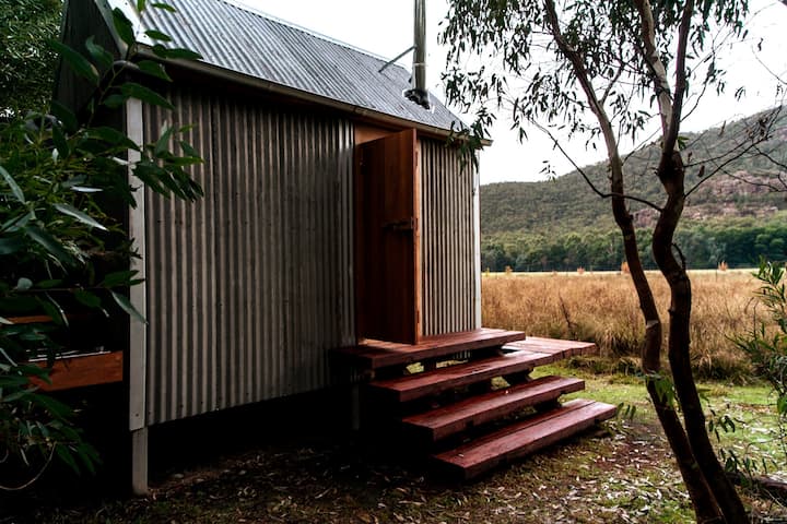  The image shows a small rustic cabin with corrugated metal siding and a pitched roof, nestled in a natural landscape. A wooden door at the top of a simple set of wooden steps leads into the cabin. Surrounding the cabin are tall grasses and a few trees, and the scene extends into a backdrop of hills and forest, providing a sense of isolation and tranquility. The overall setting feels serene, remote, and ideal for a peaceful getaway in nature.