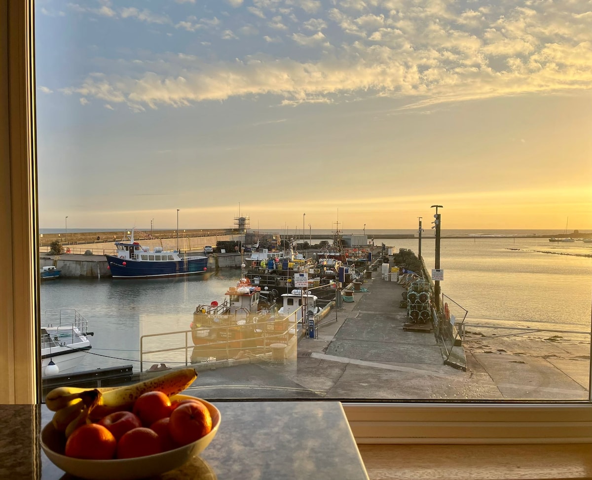 A serene view captures the working harbour at sunset, with boats docked along the pier. A bowl of fresh fruit is placed on a counter in the foreground. Soft golden light enhances the calm atmosphere reflected on the water.