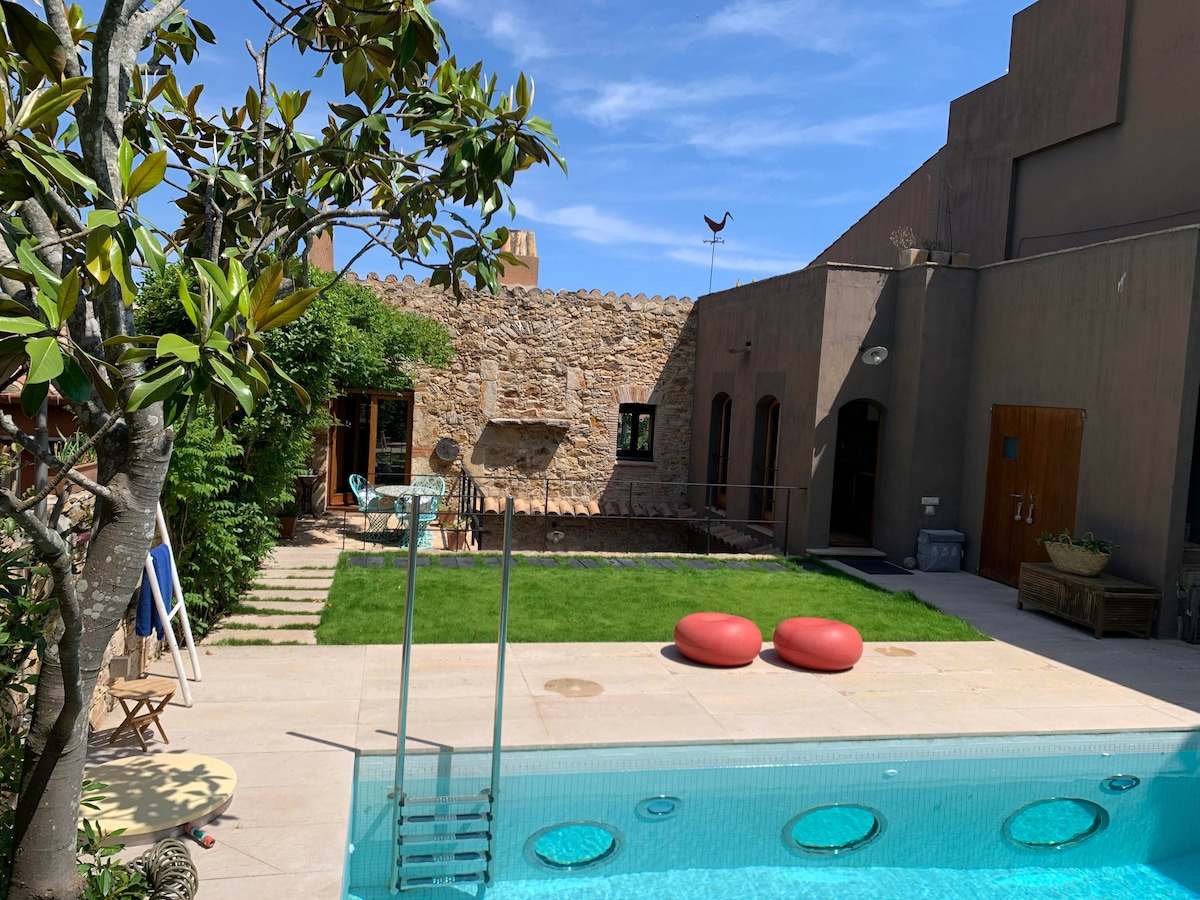 An inviting outdoor space showcases a private swimming pool in the foreground, with grassy areas bordered by stone walls. Red accent cushions are placed on the patio, complemented by a few chairs and a round table, providing a cozy setting for relaxation.
