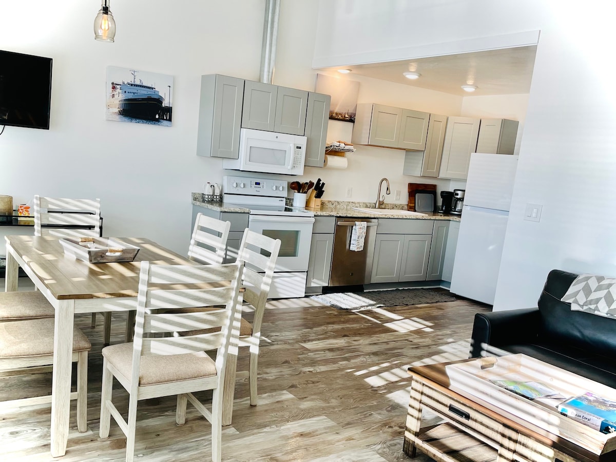An open-concept living area is displayed, featuring a dining table with six white chairs and a fully equipped kitchen visible in the background. Natural light streams through the large windows, illuminating the sleek countertops and light cabinetry. A dark sofa and coffee table are present in the space.