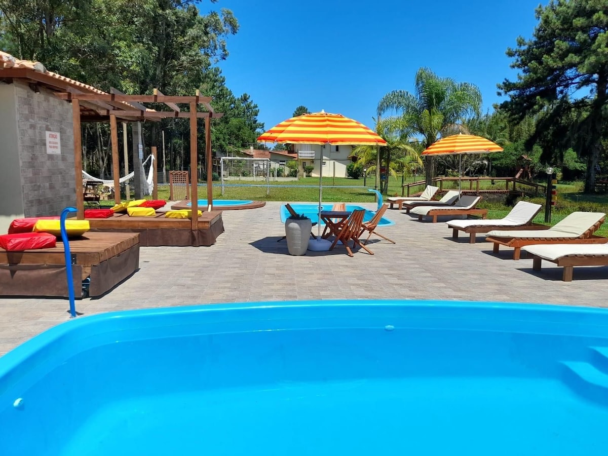 A vibrant outdoor area features a blue pool surrounded by lounge chairs and colorful umbrellas. In the background, a grassy space includes a volleyball net and shaded seating areas, inviting relaxation and recreation for guests.