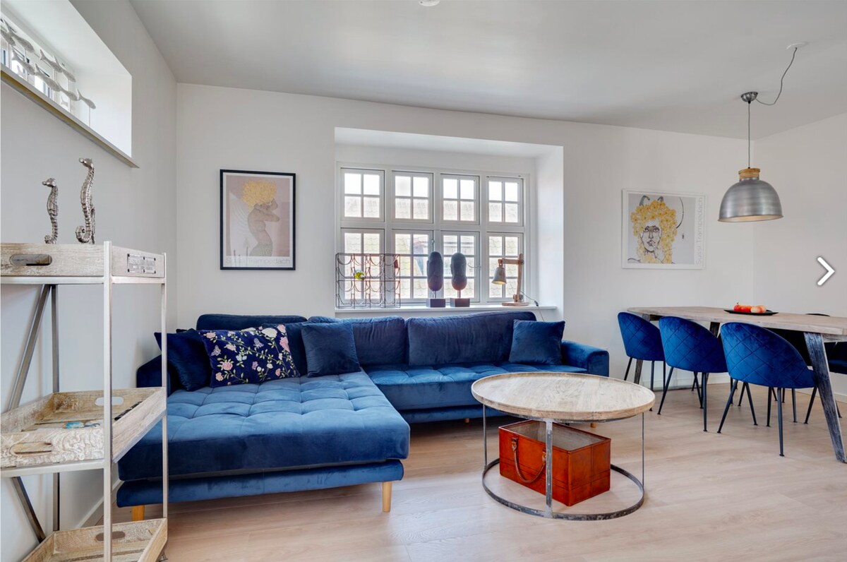 A cozy living area features a deep blue sectional sofa adorned with decorative cushions. A round marble coffee table, accompanied by a red storage box, occupies the center. Near a large window, a dining table with blue upholstered chairs is positioned, inviting gatherings.