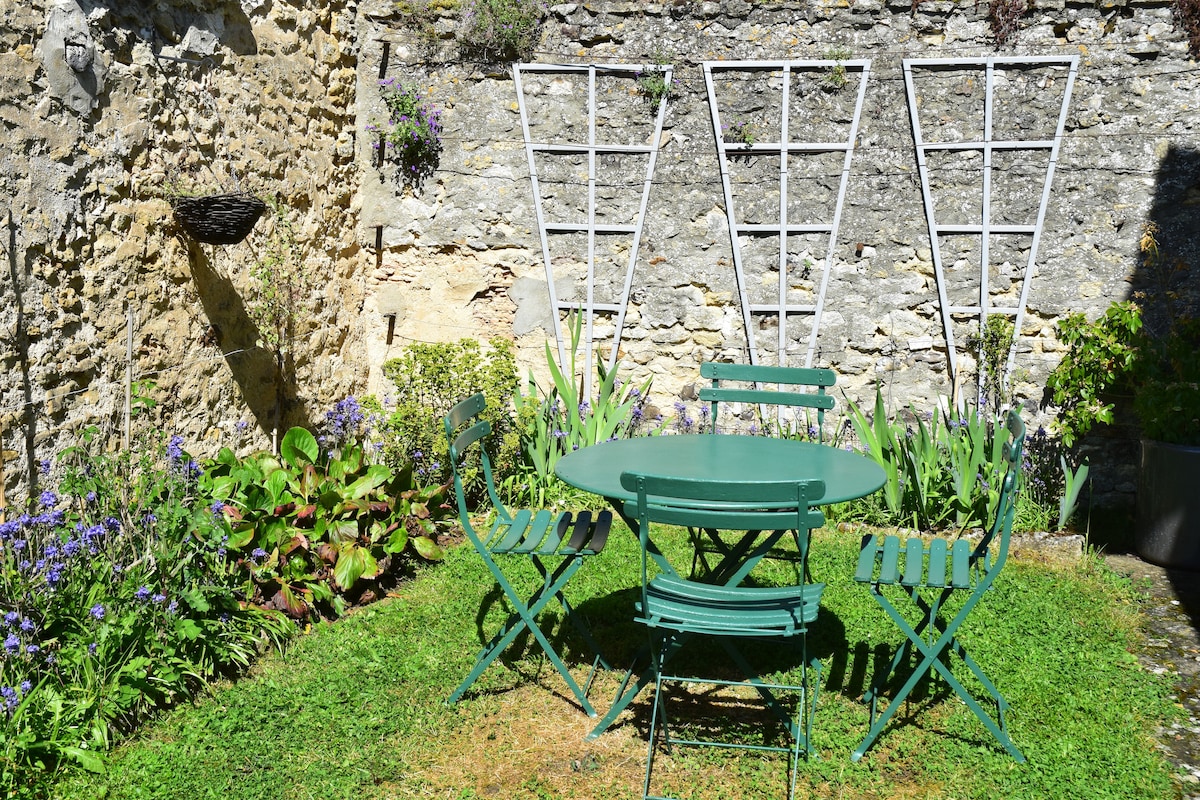 A green metal table surrounded by four matching chairs is situated on a grassy area. Lush plants and purple flowers line the stone walls, creating a serene garden space. Vertical trellises rise in the background, offering a structured contrast to the natural elements.
