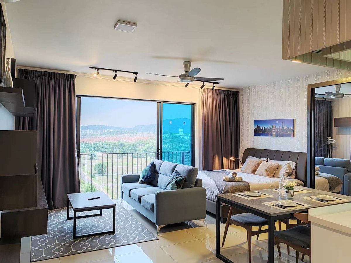 A spacious studio featuring a comfortable seating area with a three-seater sofa and a king bed. Large windows allow natural light, offering views of the surrounding landscape. A dining table and kitchenette are also visible, complemented by modern decor.
