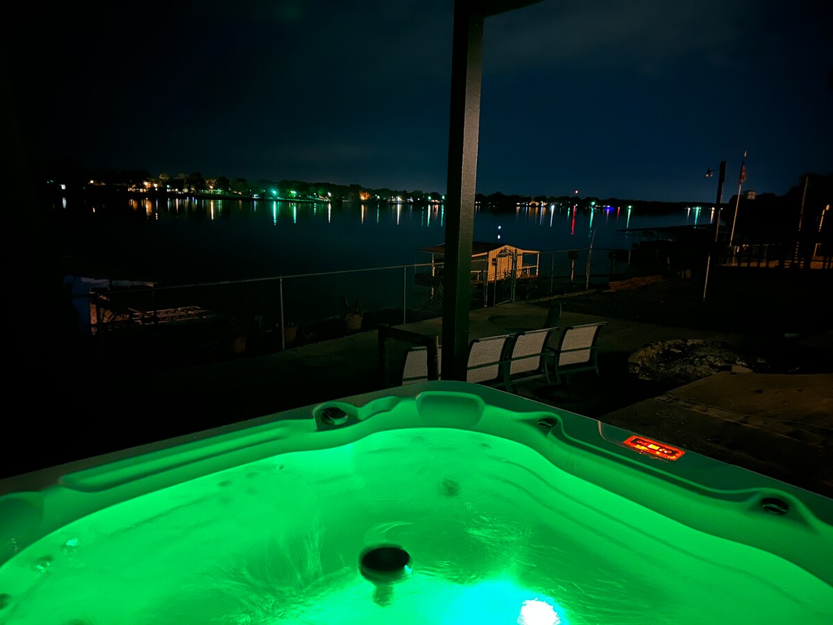 A hot tub is illuminated with soft green light, positioned on a patio facing a calm body of water. The night sky is visible, reflecting colorful lights from distant houses along the shoreline. Several outdoor chairs are arranged nearby for relaxed seating.