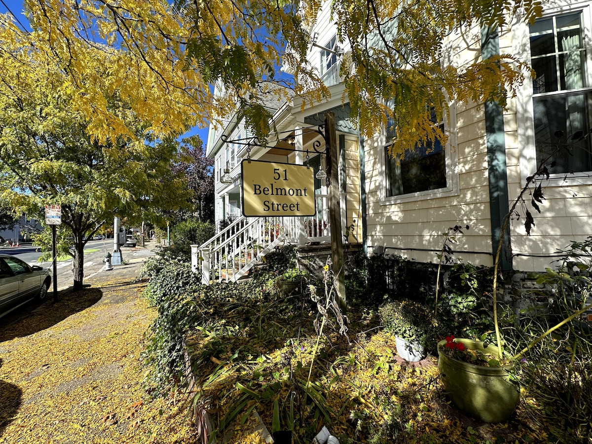 A welcoming exterior view of a two-story building features a prominent sign displaying the address '51 Belmont Street.' A lush canopy of autumn leaves frames the entrance, with a staircase leading to the main door. Bright sunlight illuminates the scene.
